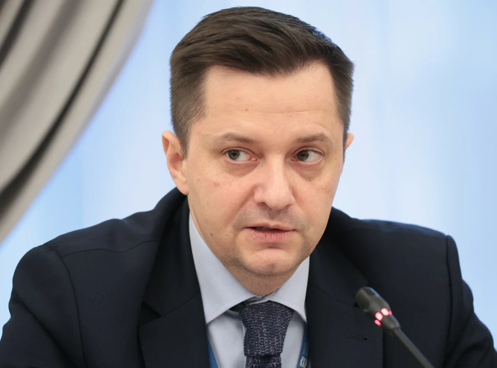 Mężczyzna w garniturze i krawacie siedzący przy mikrofonie podczas oficjalnego spotkania, tło rozmyte, po lewej stronie fragment zasłony.