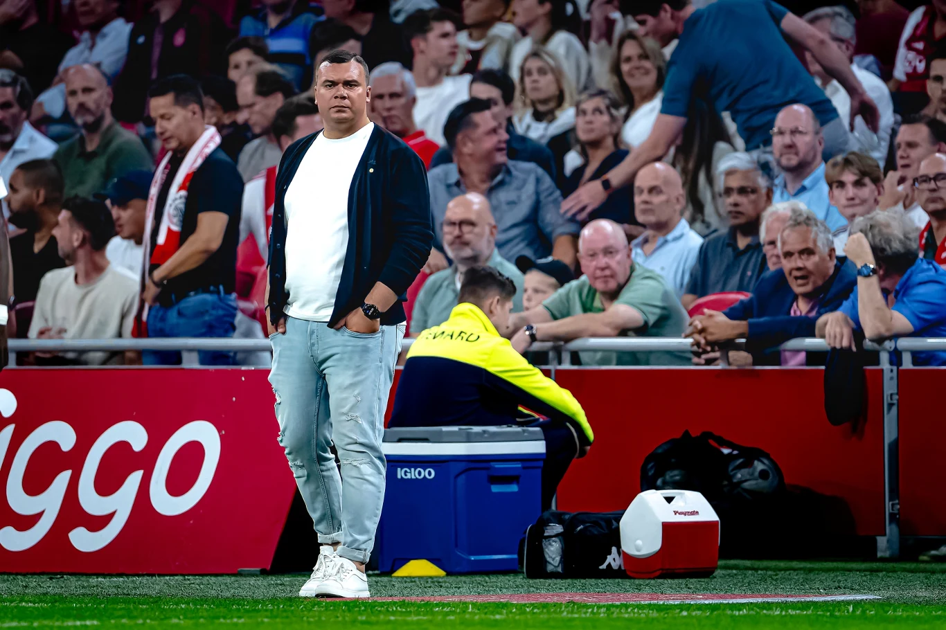 mężczyzna w jasnych spodniach i ciemnej bluzie stoi przy linii bocznej stadionu na tle siedzących kibiców; obok niego siedzi osoba w żółtej kurtce, która pochyla głowę, przy niej znajdują się chłodziarka i plecak