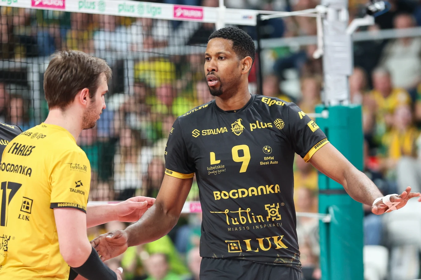 Dwóch siatkarzy w sportowych strojach podczas meczu, jeden z nich gestykuluje, na tle widać siatkę i kibiców.