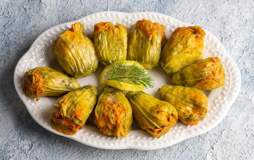Pieczone kwiaty cukinii, faszerowane ricottą, mozzarellą i orzechami włoskimi - to świetny pomysł na wykwintny obiad Talarki z kwiatów cukinii nadziewane farszem, ułożone w okrąg na białym półmisku, w centrum kawałek cytryny z gałązką koperku, całość na jasnym tle.