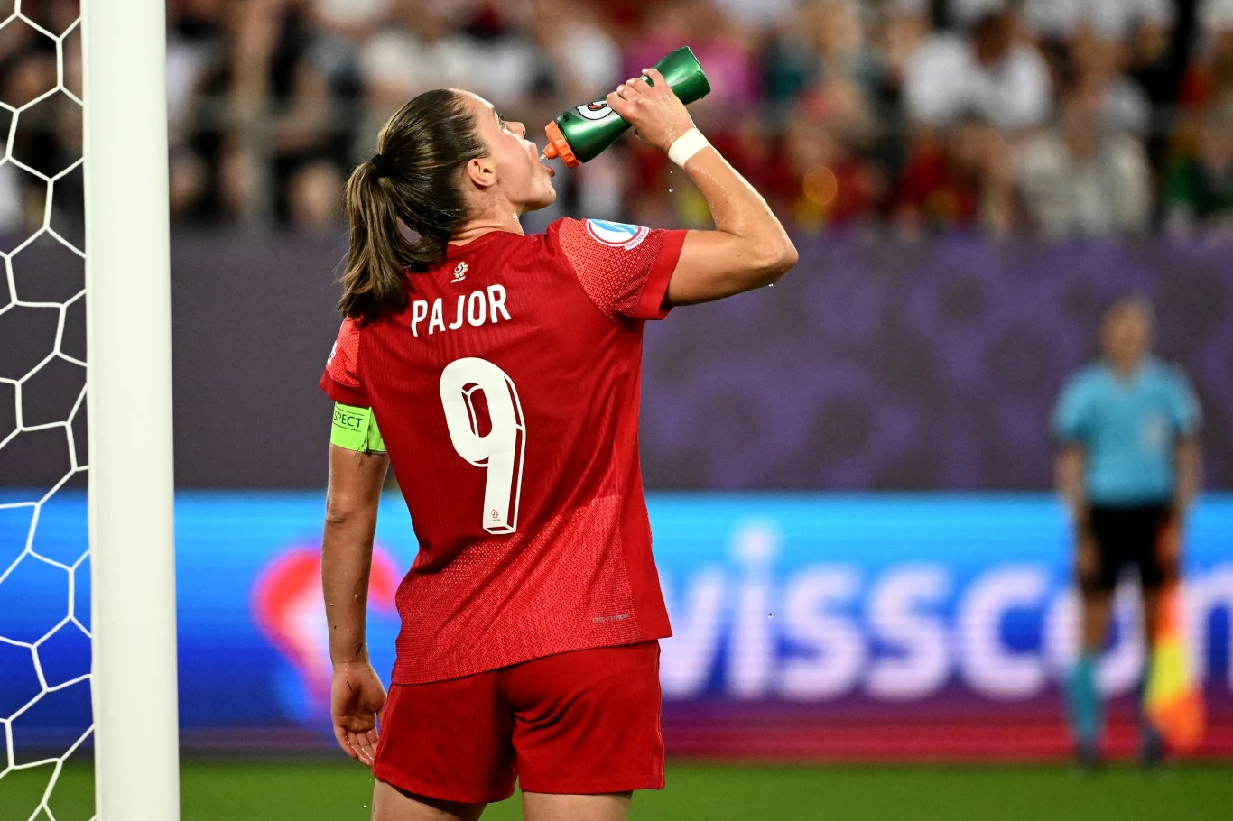 Zawodniczka w czerwonym stroju z numerem 9 i nazwiskiem Pajor pije wodę z bidonu na murawie stadionu piłkarskiego, w tle widoczni sędzia oraz rozmazana publiczność.