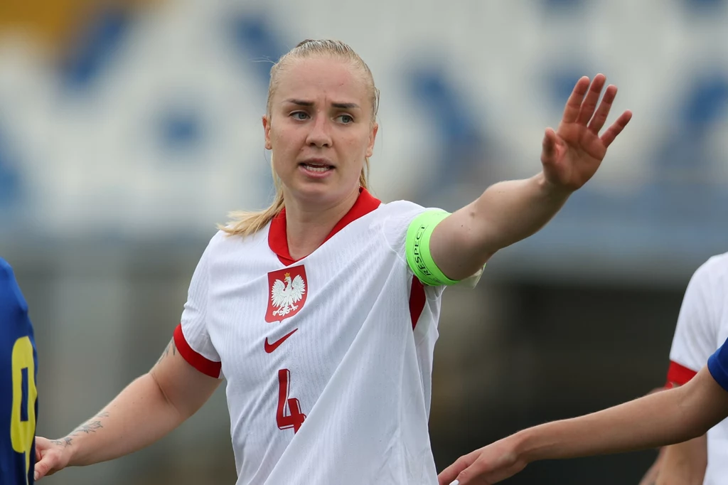 Paulina Dudek Zawodniczka w białym stroju reprezentacji Polski z opaską kapitana na ramieniu gestykuluje podczas meczu piłki nożnej, na tle rozmazanych trybun widoczna jest część innej zawodniczki w niebiesko-żółtym stroju.