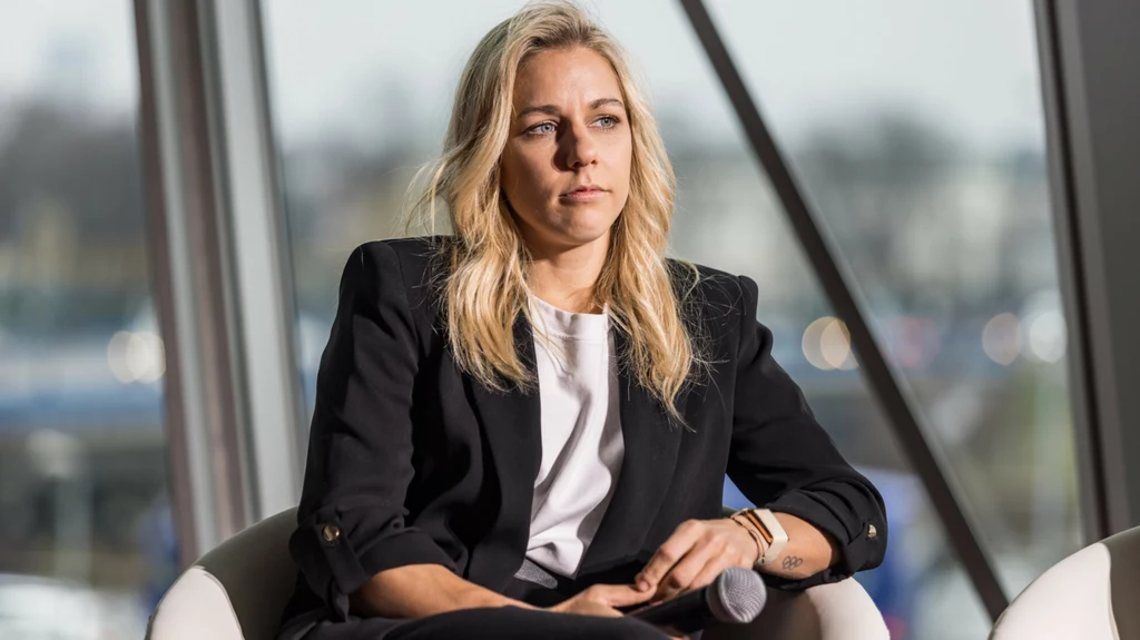 Aleksandra Mirosław Kobieta o blond włosach w ciemnej marynarce i białej bluzce, siedząca na krześle z mikrofonem w dłoni, z poważnym wyrazem twarzy w jasnym, nowoczesnym wnętrzu z dużymi oknami.