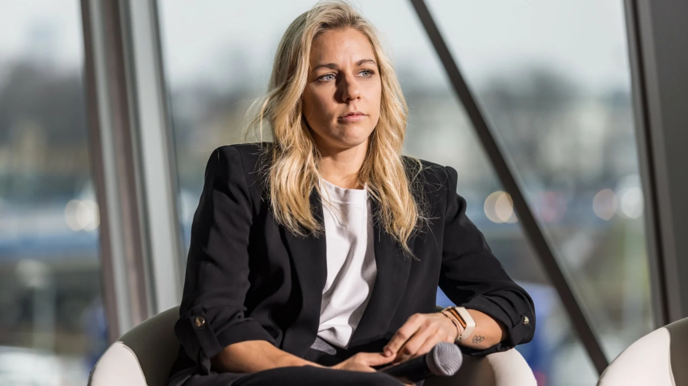 Kobieta o blond włosach w ciemnej marynarce i białej bluzce, siedząca na krześle z mikrofonem w dłoni, z poważnym wyrazem twarzy w jasnym, nowoczesnym wnętrzu z dużymi oknami.