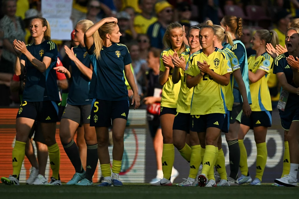 Reprezentacja Szwecji kobiet w piłce nożnej Grupa zawodniczek szwedzkiej drużyny piłkarskiej zbiera się razem na boisku, ubrane w sportowe stroje w kolorach barw narodowych, część klaska i uśmiecha się, w tle publiczność oraz elementy stadionu.