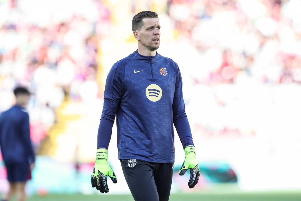 Wojciech Szczęsny Bramkarz w granatowej bluzie treningowej i jasnozielonych rękawicach na tle rozmytej publiczności stadionu piłkarskiego.