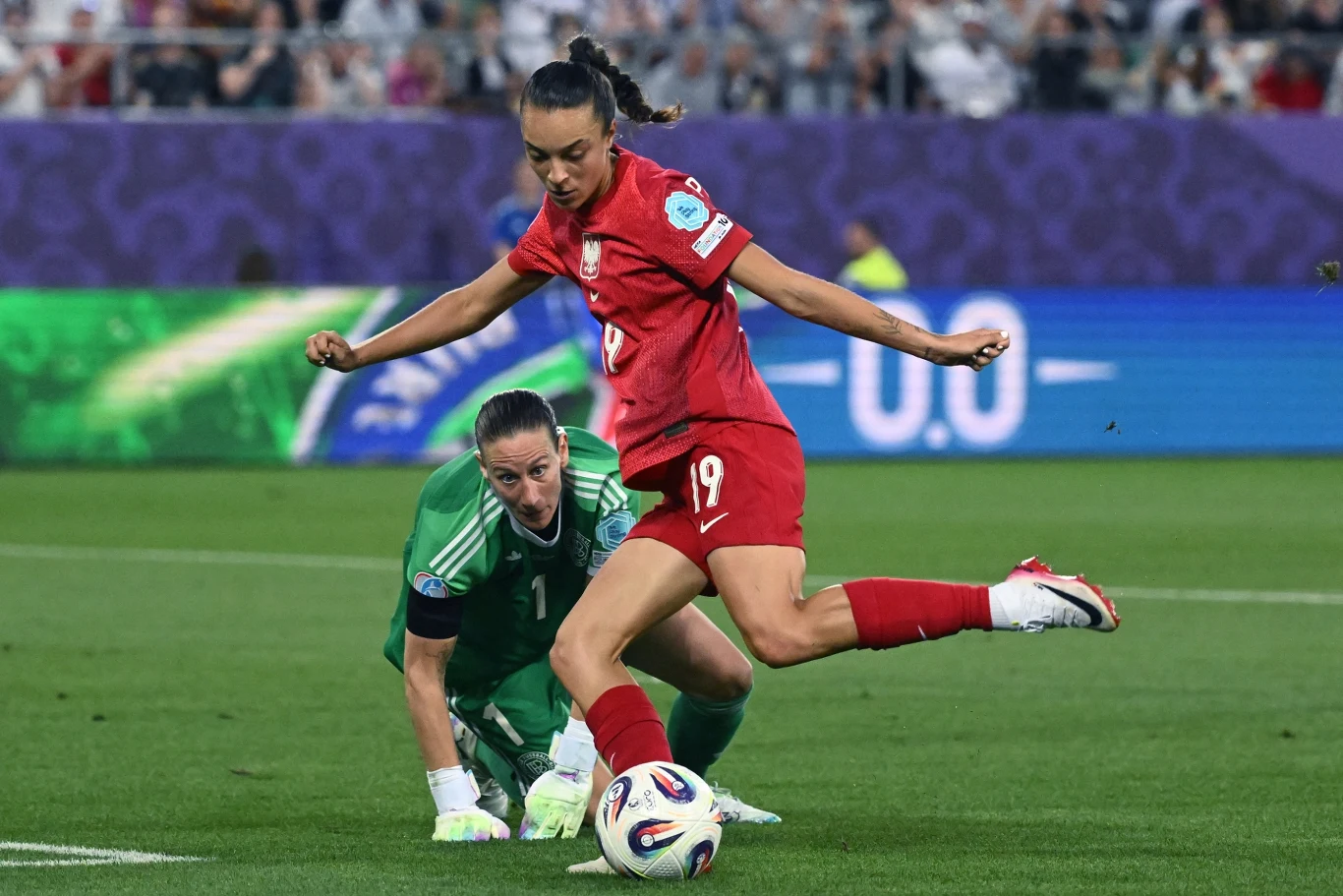 Natalia Padilla-Bidas w meczu z Niemkami Zawodniczka w czerwonym stroju piłkarskim kopie piłkę na boisku podczas meczu, przed nią klęcząca bramkarka w zielonym stroju próbująca bronić. W tle widać trybuny z kibicami.