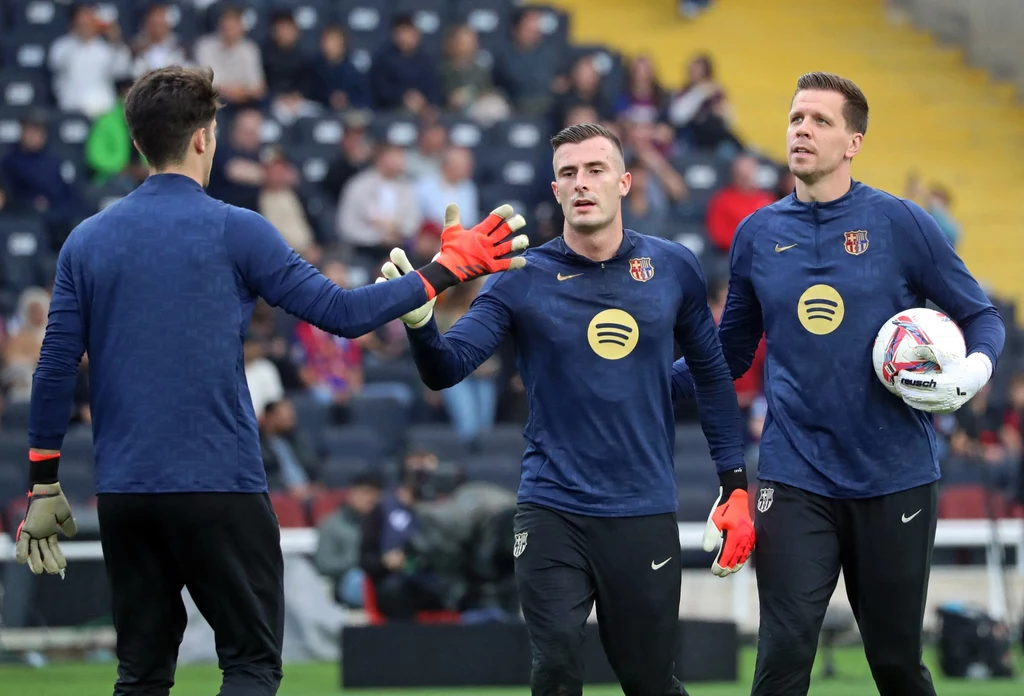 Trzech piłkarzy w sportowych strojach na stadionie, jeden z nich trzyma piłkę, wszyscy mają koszulki z logo dużej marki muzycznej, na trybunach widać widzów.