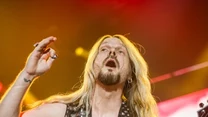 Mężczyzna z długimi blond włosami ubrany w skórzaną kamizelkę z ćwiekami gra na gitarze elektrycznej V na scenie koncertowej, w tle widoczne kolorowe światła.