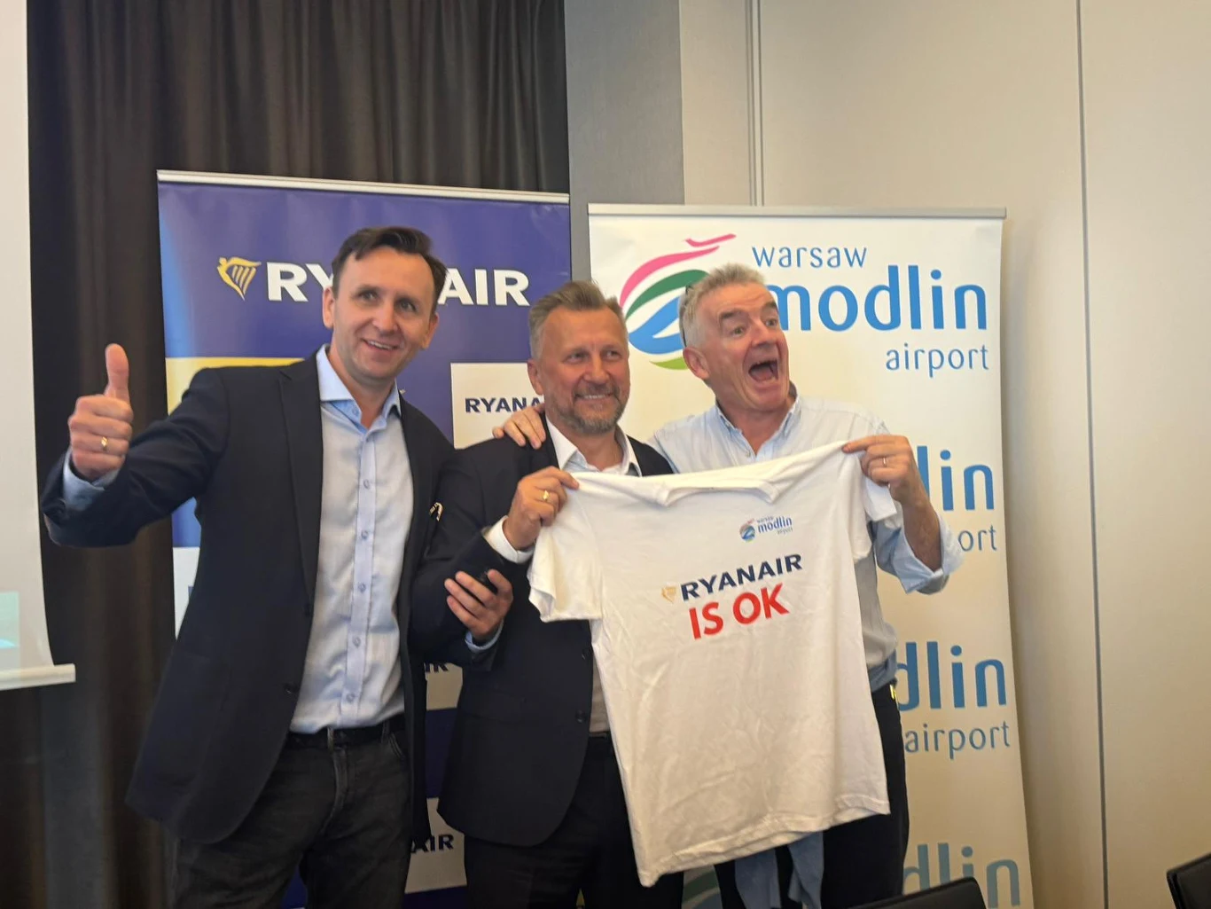 Trzech uśmiechniętych mężczyzn pozuje do zdjęcia, jeden z nich trzyma białą koszulkę z napisem 'Ryanair is OK', w tle widoczne są banery z logo Ryanair oraz warszawskiego lotniska Modlin.