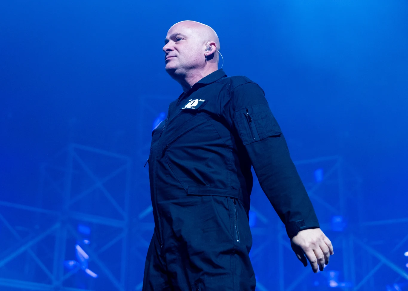 David Draiman stoi na czele grupy Disturbed Łysy mężczyzna ubrany na czarno w kombinezon stoi na scenie w mocnym, niebieskim świetle, w tle widoczne są elementy konstrukcji oświetleniowej sceny.