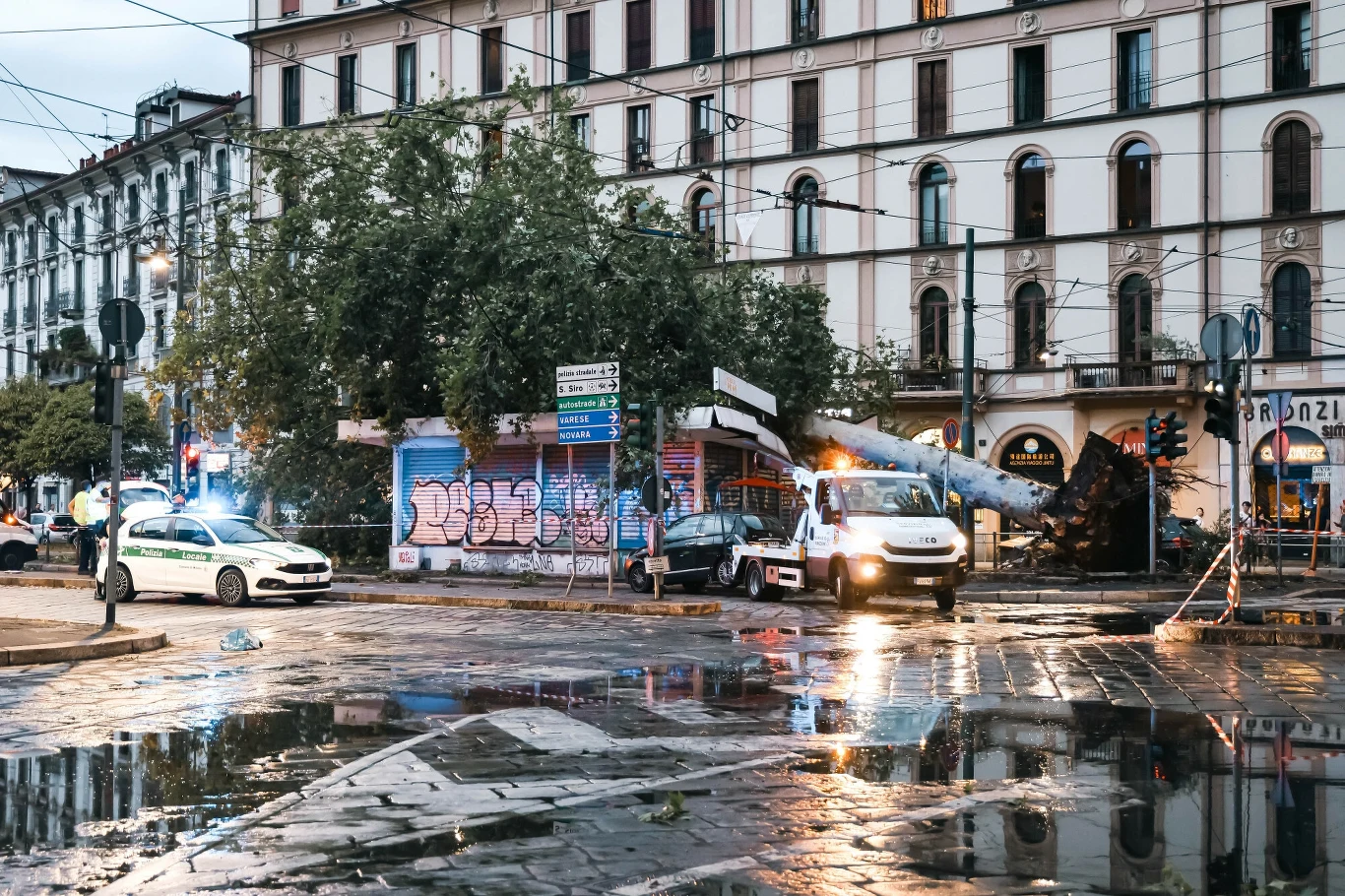 Skutki przejścia gwałtownej burzy przez Mediolan. Potężne wichury i ulewy oprócz Włoch doświadczyły również Węgry i Słowację Przewrócone drzewo leży na budynku na ruchliwym miejskim skrzyżowaniu po deszczu; służby ratownicze zabezpieczają teren, a po mokrej nawierzchni jezdni odbija się światło policyjnych i ratunkowych pojazdów.