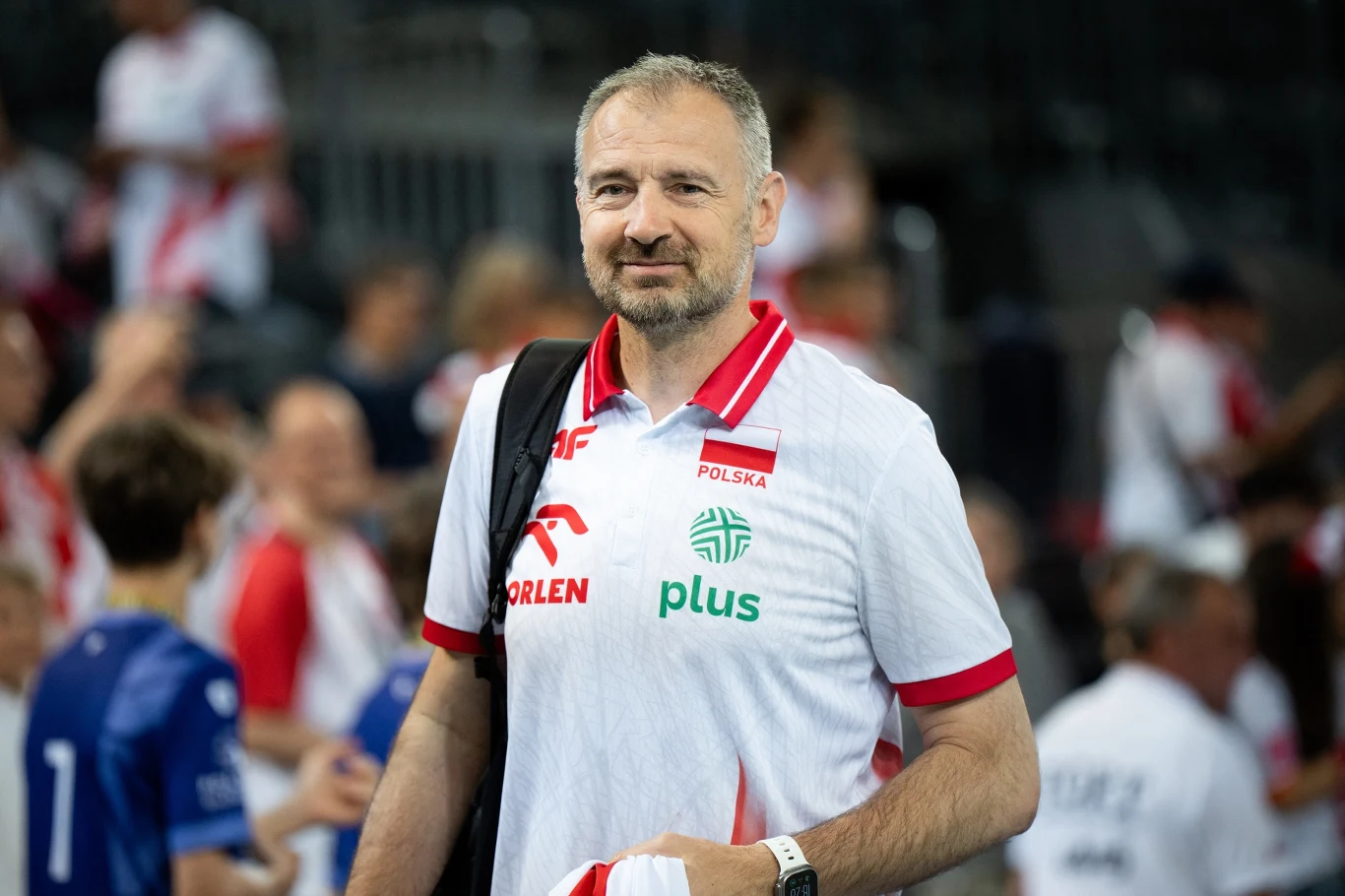 Mężczyzna z zarostem w sportowej koszulce z logotypami sponsorów, z torbą na ramieniu i ręcznikiem, stoi na tle rozmytego tłumu ubranych w sportową odzież.