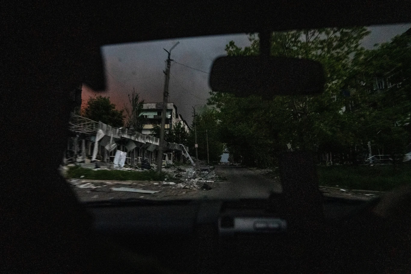 Ruiny budynków oraz zniszczona ulica widziana z wnętrza samochodu, ciemna atmosfera oraz ślady po zniszczeniach świadczą o niedawnych działaniach wojennych lub katastrofie.