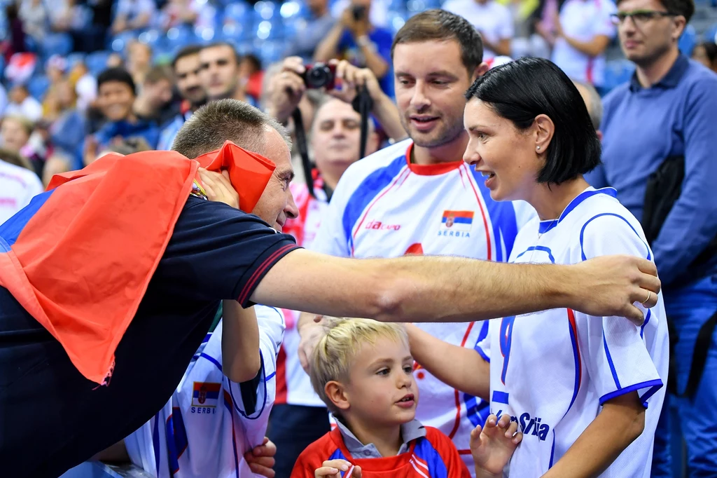 Grupa kibiców ubranych w stroje z flagą Serbii rozmawia i celebruje na trybunach podczas wydarzenia sportowego, w centrum uwagi znajdują się uśmiechnięci dorośli oraz dziecko otoczeni tłumem sympatyków.