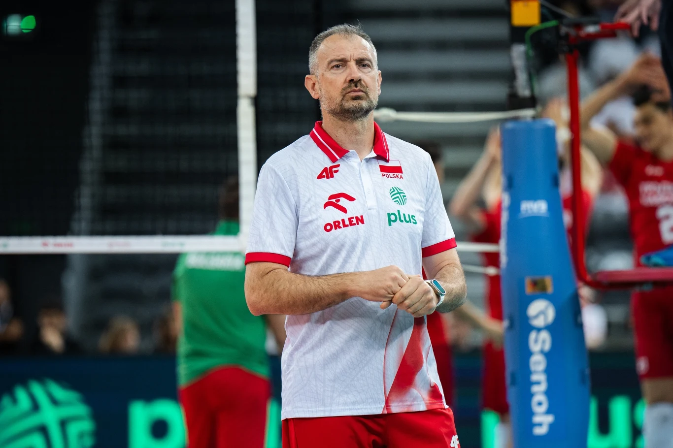 Nikola Grbić Trener siatkarski w białej koszulce z logotypami sponsorów stoi na hali sportowej, w tle zawodnicy w czerwonych strojach oraz siatka i elementy boiska do siatkówki.