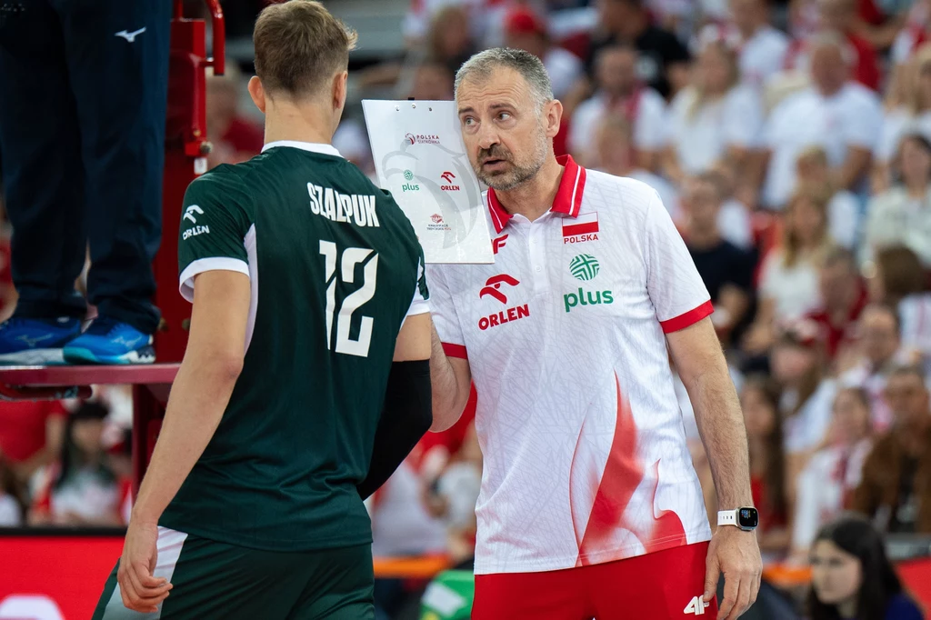 Trener reprezentacji Polski w białej koszulce rozmawia z siatkarzem w ciemnym stroju podczas meczu siatkówki, tło wypełniają kibice na trybunach.