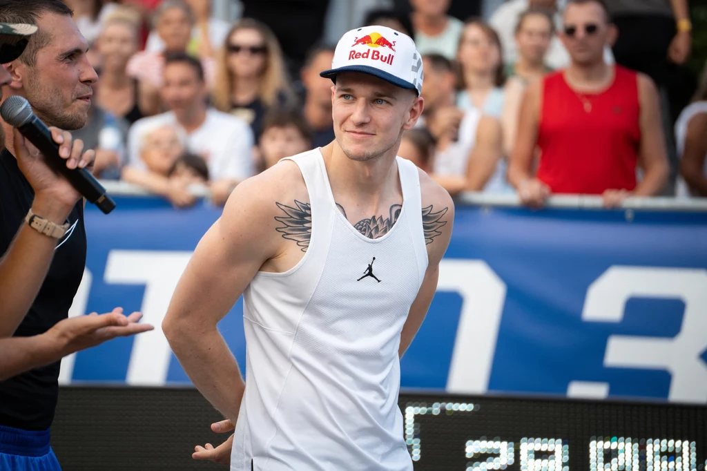 Piotr "Grabo" Grabowski Młody mężczyzna w białej koszulce na ramiączkach i czapce z logo Red Bull, z widocznymi tatuażami na ramionach, stoi podczas wydarzenia sportowego, wokół znajduje się tłum ludzi i prowadzący rozmawiający przez mikrofon.