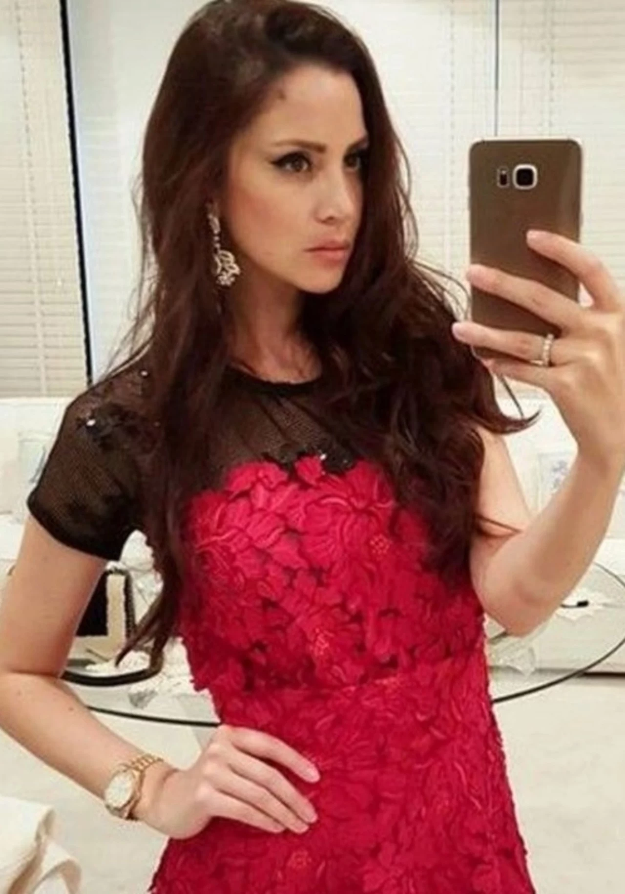 Młoda kobieta z długimi, ciemnobrązowymi włosami robi selfie w lustrze. Ubrana jest w elegancką, czerwoną sukienkę z koronkowymi aplikacjami i czarną górą oraz efektowne kolczyki. W tle widać jasny, nowoczesny wystrój wnętrza.