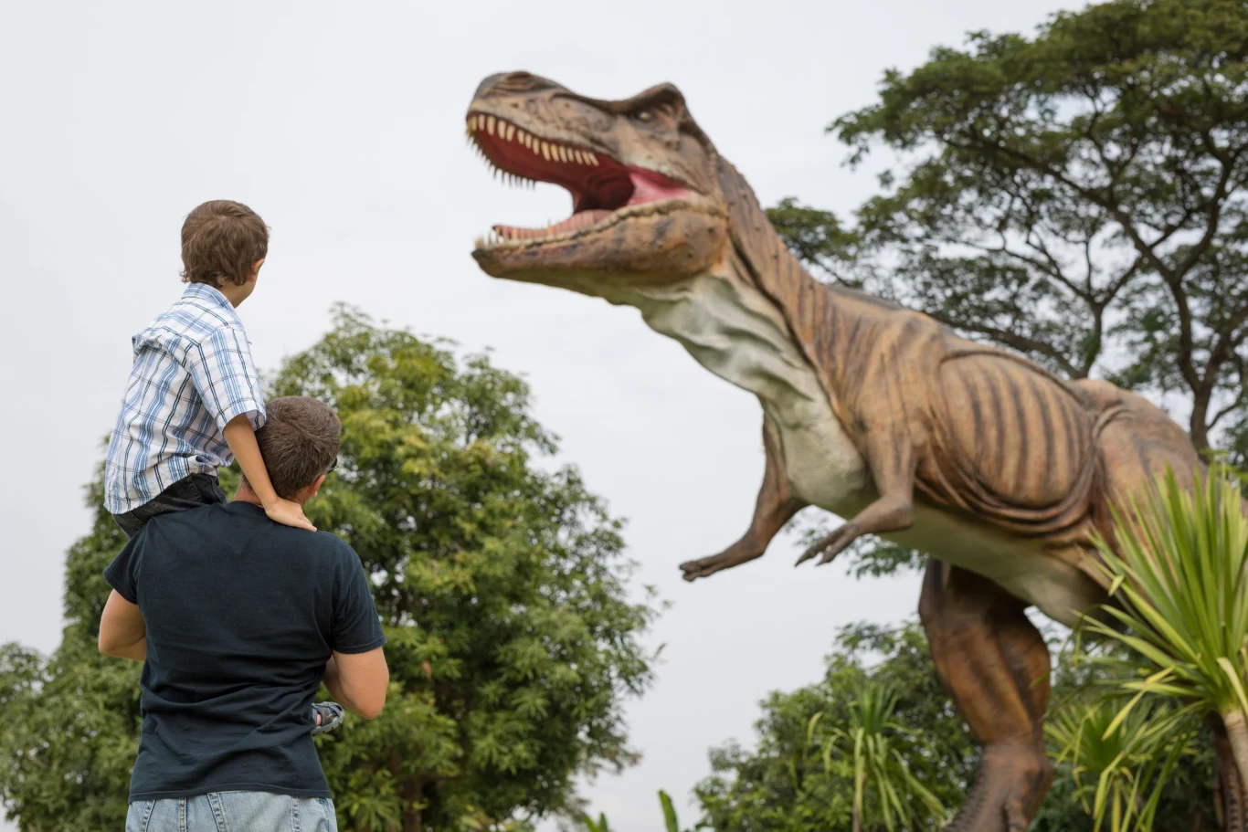 Mężczyzna trzyma chłopca na ramionach, obaj patrzą na dużą, realistyczną figurę dinozaura typu tyranozaur w otoczeniu zieleni i drzew w parku lub ogrodzie tematycznym.