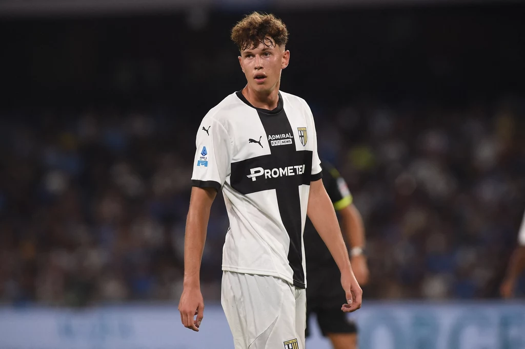 Mateusz Kowalski w barwach Parmy Piłkarz w białej koszulce z czarnymi paskami na stadionie podczas meczu, w tle rozmazane sylwetki innych graczy.