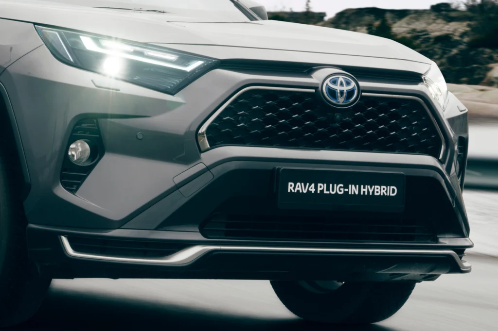 Przedni fragment szarego samochodu Toyota RAV4 Plug-in Hybrid z wyraźnie widoczną atrapą chłodnicy, reflektorami LED oraz tabliczką modelu na tle rozmytego krajobrazu.