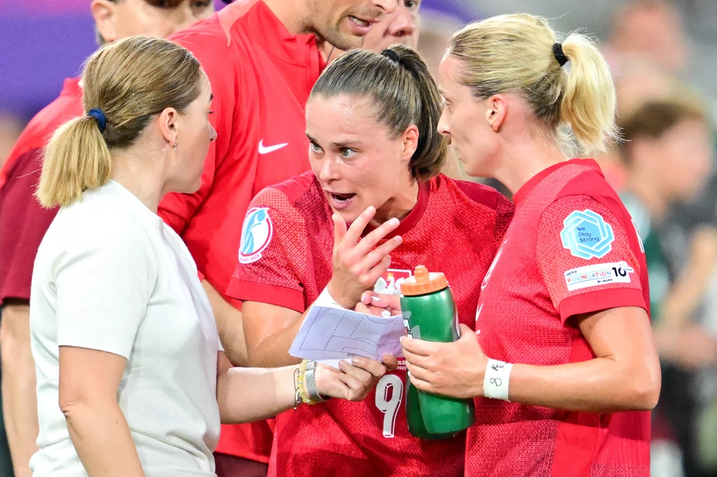 Nina Patalon i Ewa Pajor podczas Euro kobiet 2025 Dwie piłkarki w czerwonych koszulkach i sztab trenerski intensywnie omawiają taktykę, jedna z zawodniczek trzyma notatkę, druga bidon, pełne emocji twarze wskazują na ważny moment sportowego spotkania.