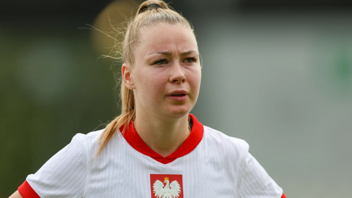 Piłkarka w białej koszulce z orłem na piersi, z włosami związanymi w kucyk, stoi na boisku, patrząc przed siebie z poważnym wyrazem twarzy.