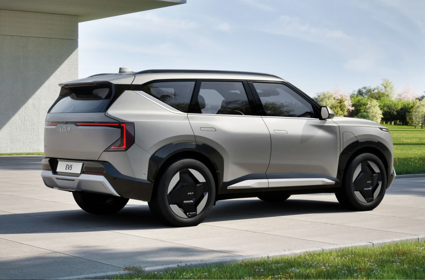 Nowoczesny, srebrny samochód SUV marki Kia EV9 zaparkowany na podjeździe w otoczeniu zieleni i nowoczesnej architektury, widoczny z tyłu pod lekkim kątem.