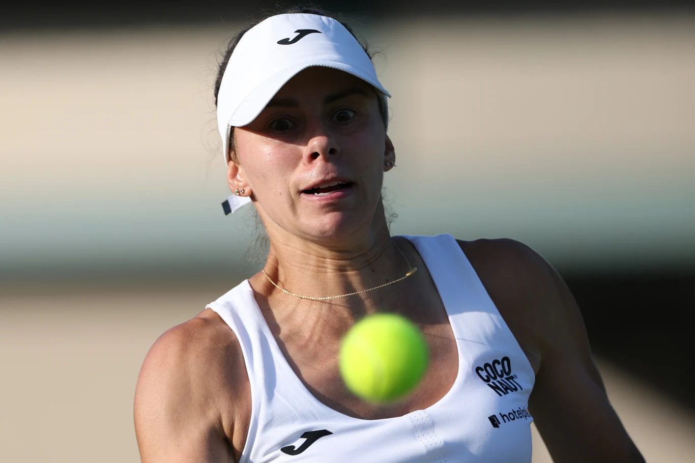 Tenisistka w białym stroju sportowym podczas intensywnej gry śledzi wzrokiem lecącą piłkę tenisową, na twarzy skupienie i determinacja, w tle rozmyte boisko.