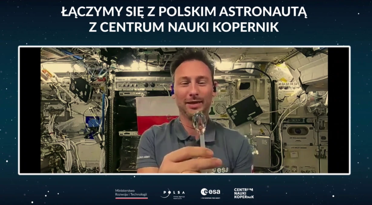 Sławosz Uznański-Wiśniewski przeprowadza jeden z eksperymentów. Tak zachowuje się woda na ISS Polski astronauta na tle wnętrza Międzynarodowej Stacji Kosmicznej prezentuje przedmiot, za nim widoczna polska flaga oraz skomplikowane urządzenia stacji kosmicznej.