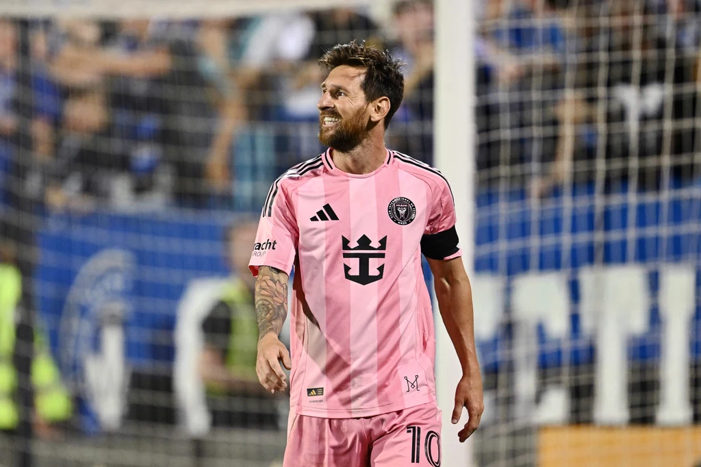 Lionel Messi Piłkarz w różowym stroju drużyny Inter Miami CF znajduje się na murawie przed bramką, podczas meczu piłkarskiego, uśmiecha się, w tle rozmyta publiczność i siatka bramki.