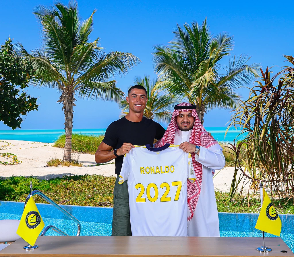 Cristiano Ronaldo przedłużył kontrakt z Al Nassr Dwie osoby stojące obok siebie, jedna z nich trzyma koszulkę z nazwiskiem Ronaldo i liczbą 2027, w tle widoczny tropikalny krajobraz z palmami, basenem i plażą oraz jasnoniebieskim morzem, na stole przed nimi znajdują się dwa żółte proporczyki.