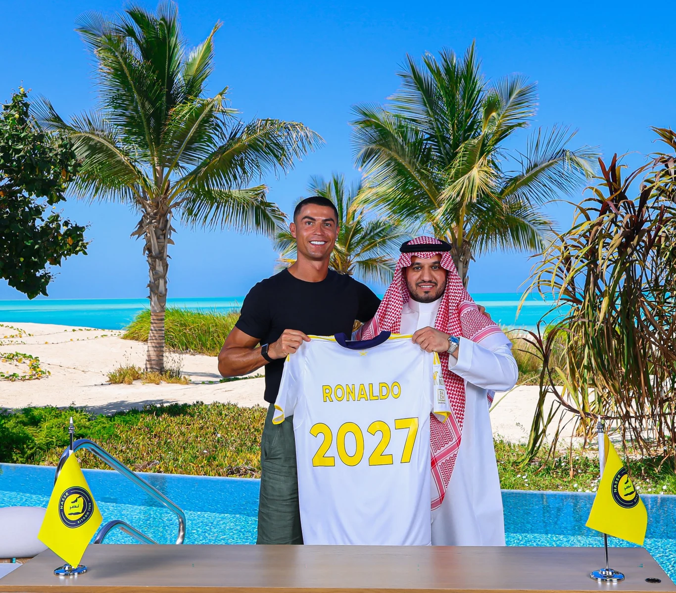 Dwie osoby stojące obok siebie, jedna z nich trzyma koszulkę z nazwiskiem Ronaldo i liczbą 2027, w tle widoczny tropikalny krajobraz z palmami, basenem i plażą oraz jasnoniebieskim morzem, na stole przed nimi znajdują się dwa żółte proporczyki.