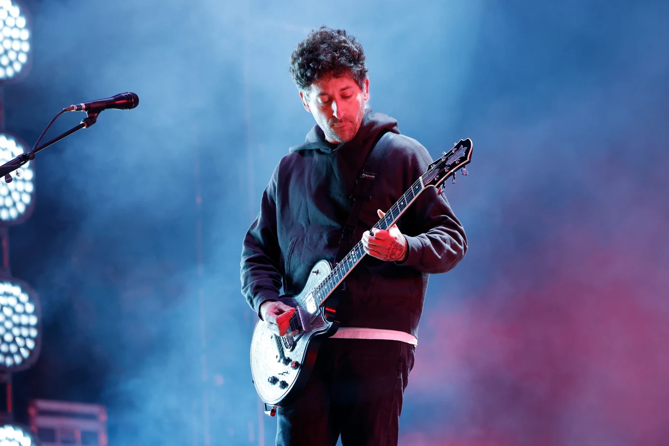 Joe Trohman - gitarzysta Fall Out Boy Mężczyzna grający na gitarze elektrycznej podczas koncertu, otoczony kolorowym oświetleniem i dymem scenicznym, skierowany wzrok w stronę instrumentu.