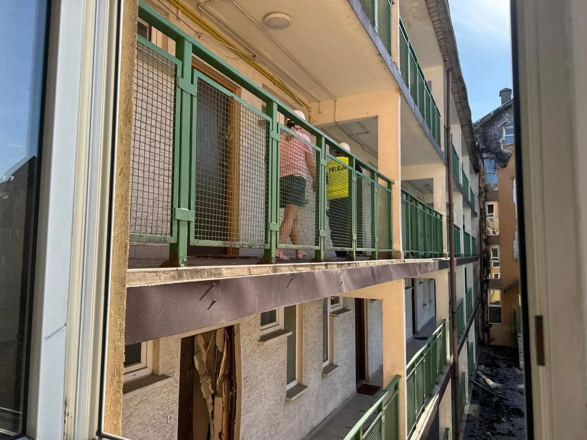 Skutki pożaru kompleksu mieszkalnego w Ząbkach Osiedlowy korytarz na piętrze kamienicy z zielonymi balustradami, przez które widać osobę idącą z żółtym wiadrem, na dolnej kondygnacji widoczne uszkodzenia drzwi oraz ściany.