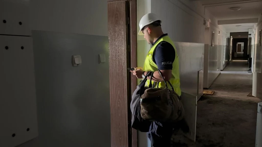 Skutki pożaru kompleksu mieszkalnego w Ząbkach Policjant w kamizelce odblaskowej i kasku ochronnym stoi w opuszczonym lub remontowanym budynku, trzymając dużą torbę i patrząc na ekran telefonu, w tle korytarz o zniszczonej podłodze
