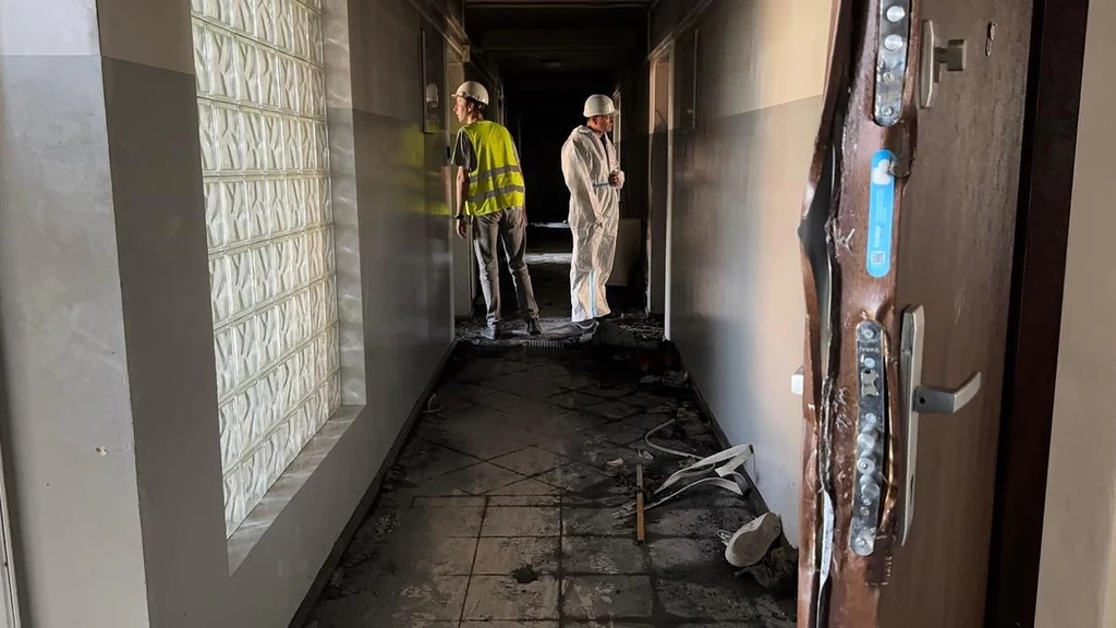 Spalony korytarz budynku z widocznymi śladami ognia i sadzy, przed którym stoją dwaj mężczyźni w odzieży ochronnej i kaskach, sprawdzający stan pomieszczeń.