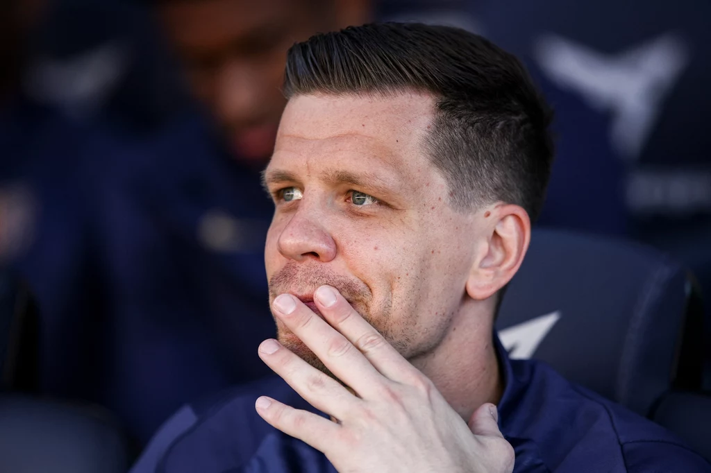 Wojciech Szczęsny Mężczyzna z zarostem, ubrany w granatową kurtkę, siedzi zamyślony i opiera dłoń na twarzy, w tle rozmyte postaci i ciemne tło.
