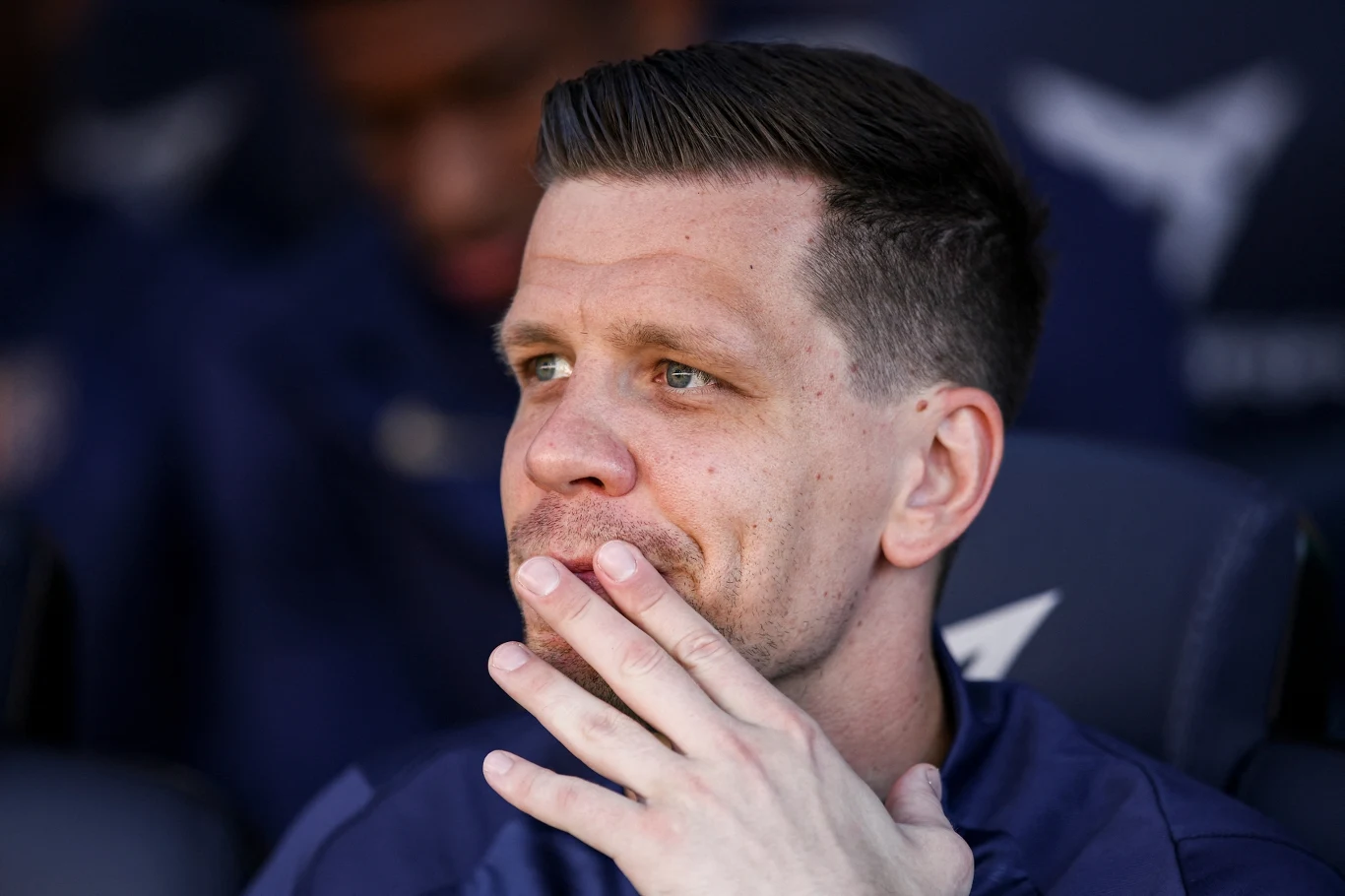 Mężczyzna z zarostem, ubrany w granatową kurtkę, siedzi zamyślony i opiera dłoń na twarzy, w tle rozmyte postaci i ciemne tło.