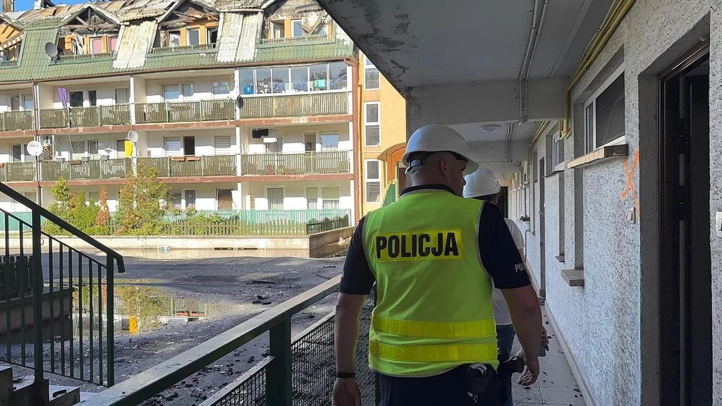 Funkcjonariusz policji w żółtej kamizelce z napisem 'Policja', w kasku ochronnym, idzie korytarzem uszkodzonego przez pożar budynku. Na zewnątrz widoczny jest wielokondygnacyjny blok z wyraźnymi śladami zniszczeń, spalonym dachem oraz balkonami z osmol...