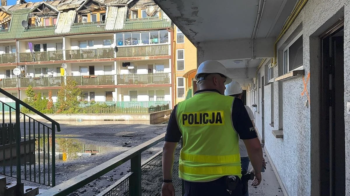 Policja opublikowała zdjęcia ukazujące skutki pożaru w Ząbkach Funkcjonariusz policji w żółtej kamizelce z napisem 'Policja', w kasku ochronnym, idzie korytarzem uszkodzonego przez pożar budynku. Na zewnątrz widoczny jest wielokondygnacyjny blok z wyraźnymi śladami zniszczeń, spalonym dachem oraz balkonami z osmol...