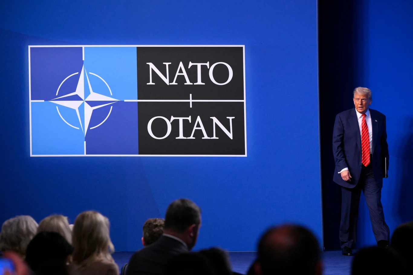 Donald Trump wychodzi na scenę podczas konferencji lub spotkania NATO, w tle duże logo NATO na niebieskim tle, zebrana publiczność siedzi przodem do sceny.