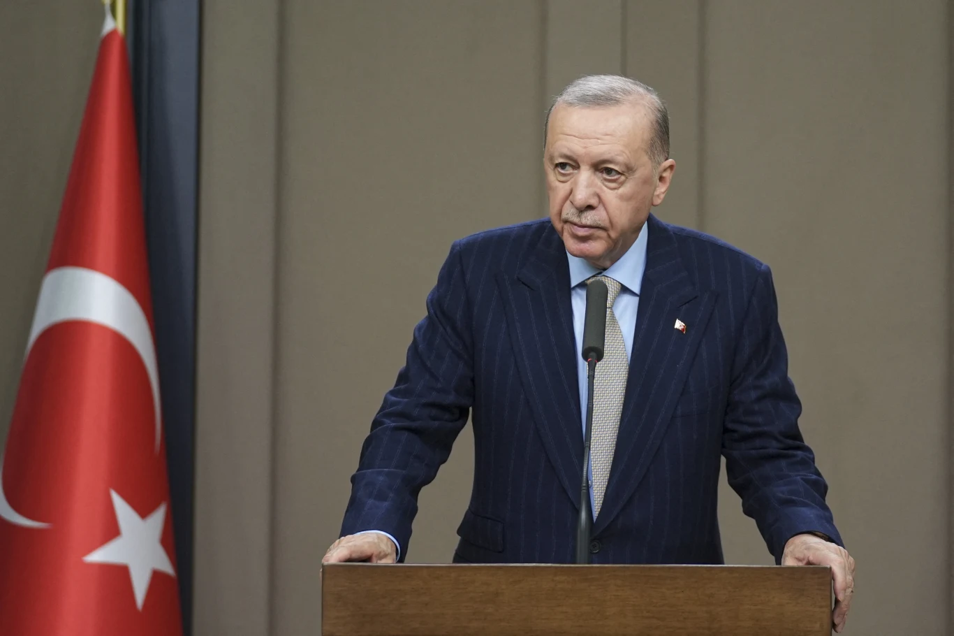 Tureckie służby aresztowały kolejnych przeciwników politycznych prezydenta Recepa Tayyipa Erdogana Mężczyzna w garniturze stojący za mównicą, przemawia do mikrofonu, po lewej stronie kadr flagi Turcji, tło jednolite, formalna atmosfera.