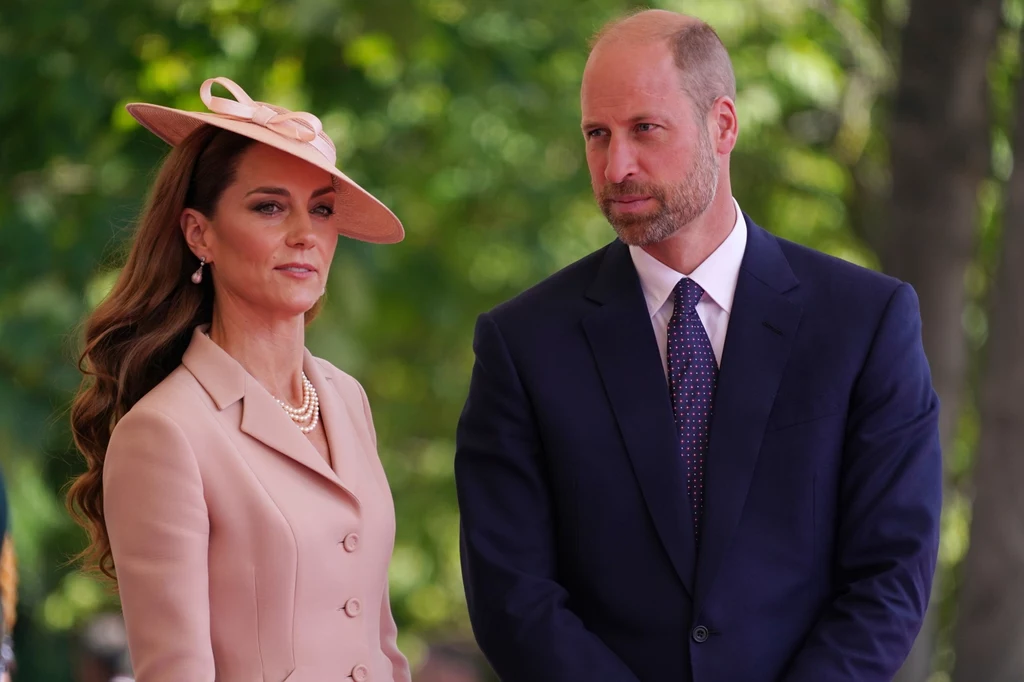 Kilka dni po wizycie w szpitalu Kate towarzyszyła podczas ważnej uroczystości mężowi Elegancko ubrana kobieta w jasnoróżowym kostiumie i kapeluszu stoi obok mężczyzny w ciemnym garniturze na tle zieleni, oboje wyglądają poważnie i skupieni.