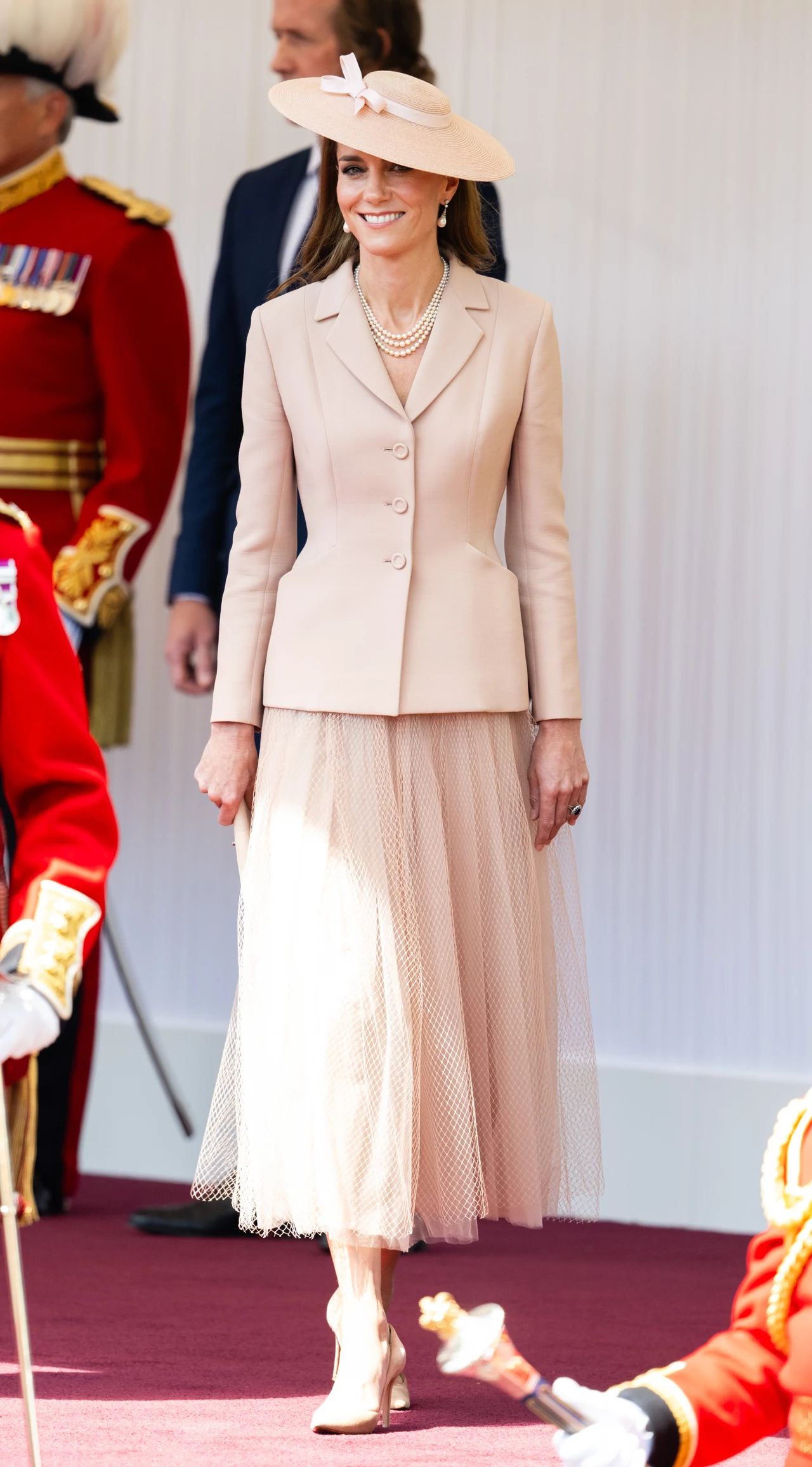 Stylizacja księżnej Kate nawiązywała do francuskiej mody Elegancka kobieta w jasnym kostiumie i kapeluszu spaceruje po czerwonym dywanie, wokół niej widoczni są żołnierze w czerwonych mundurach paradnych.