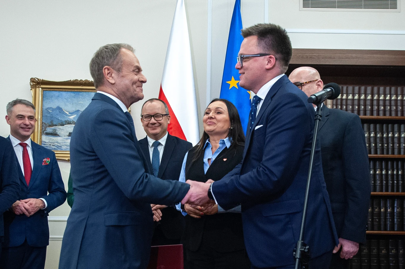 Premier Donald Tusk i marszałek Sejmu Szymon Hołownia Grupa polityków w eleganckich garniturach w oficjalnym pomieszczeniu, dwóch mężczyzn w centrum ściska sobie dłonie, w tle widoczne flagi Polski i Unii Europejskiej oraz półki z książkami.