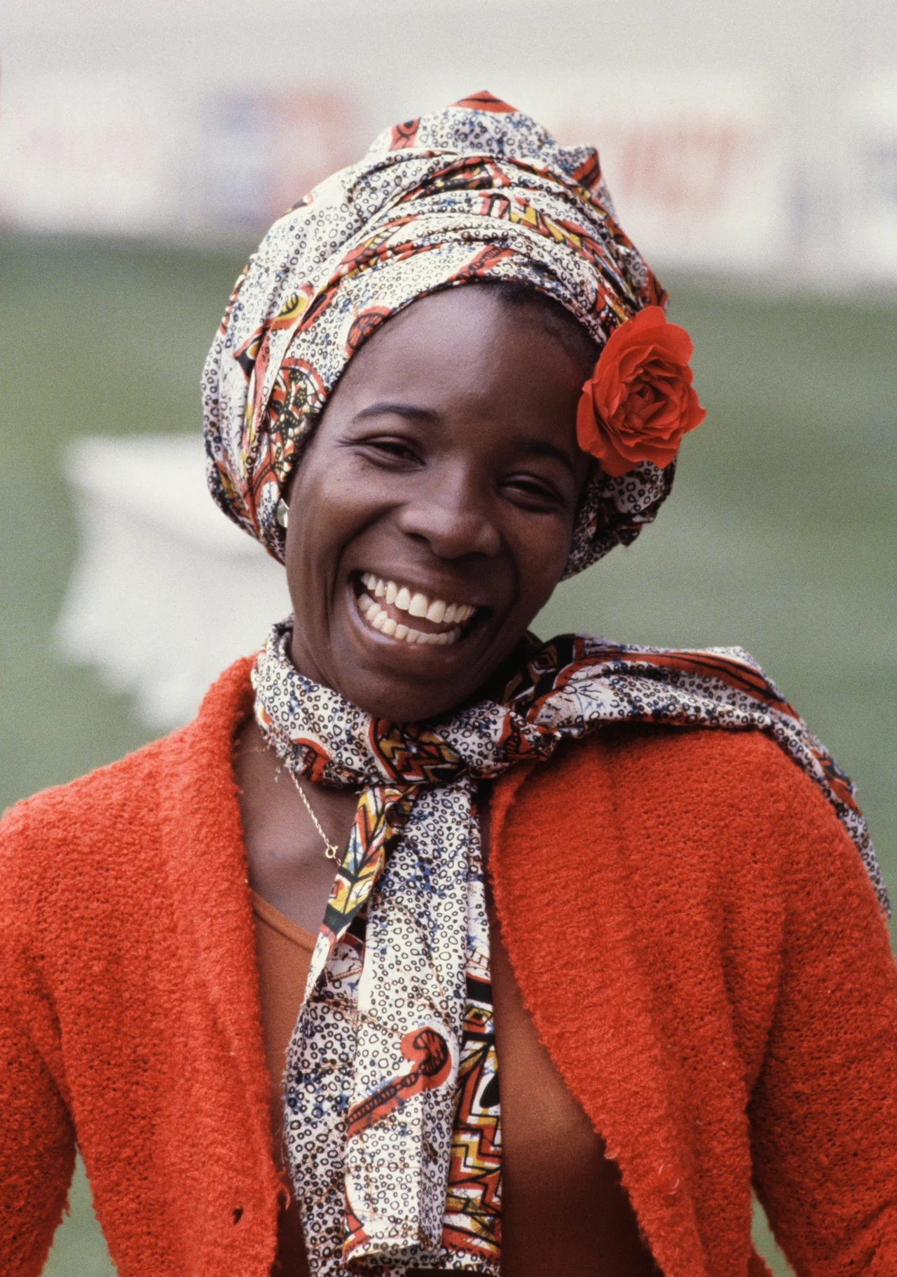 Rita Marley w Paryżu - 1 maja 1980 r. Uśmiechnięta kobieta w kolorowym stroju, ozdobiona czerwoną różą w chustce na głowie i ubrana w czerwony sweter, stoi na tle zielonego trawnika.