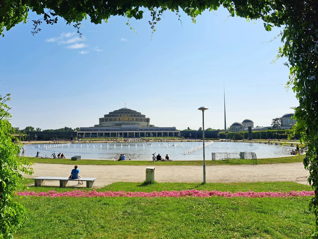 Hala Stulecia we Wrocławiu Pergola z widokiem na Halę Stulecia we Wrocławiu, centralnie duży okrągły staw otoczony ścieżkami i zielenią, w tle charakterystyczny budynek hali, wokół rozproszeni spacerowicze i ławki, całość w słoneczny dzień.