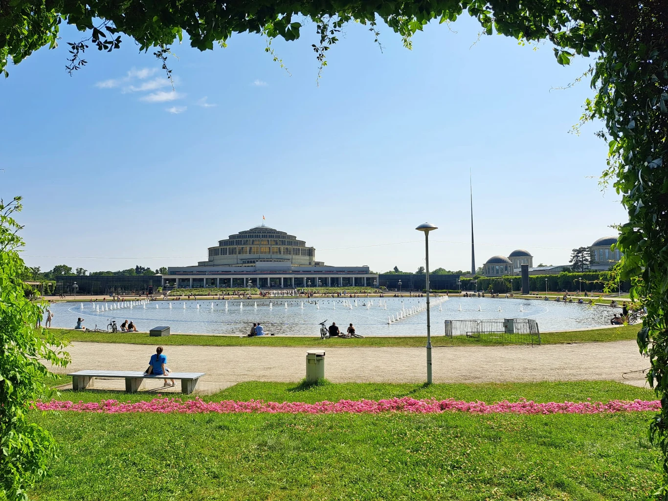 Hala Stulecia we Wrocławiu Pergola z widokiem na Halę Stulecia we Wrocławiu, centralnie duży okrągły staw otoczony ścieżkami i zielenią, w tle charakterystyczny budynek hali, wokół rozproszeni spacerowicze i ławki, całość w słoneczny dzień.